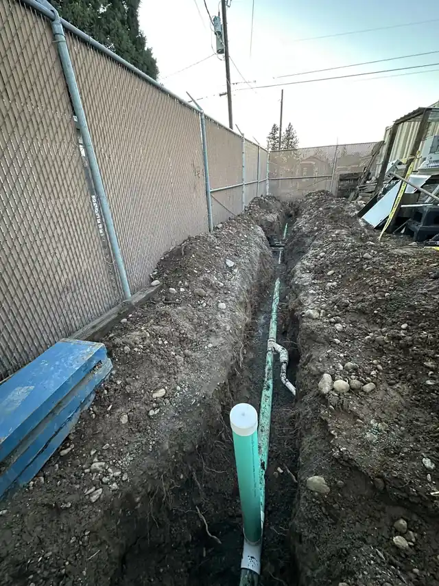 An open sewer trench with shoring panels along a fence line, opened after the customer approved the repair