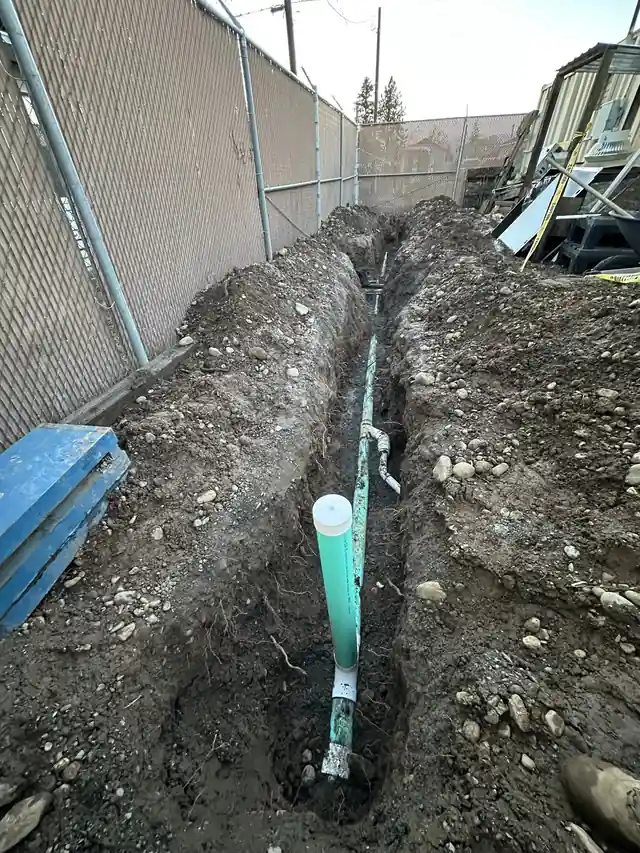 New green PVC sewer line and a white cleanout riser in an open trench along a chain-link fence, dug by CSR Plumbing
