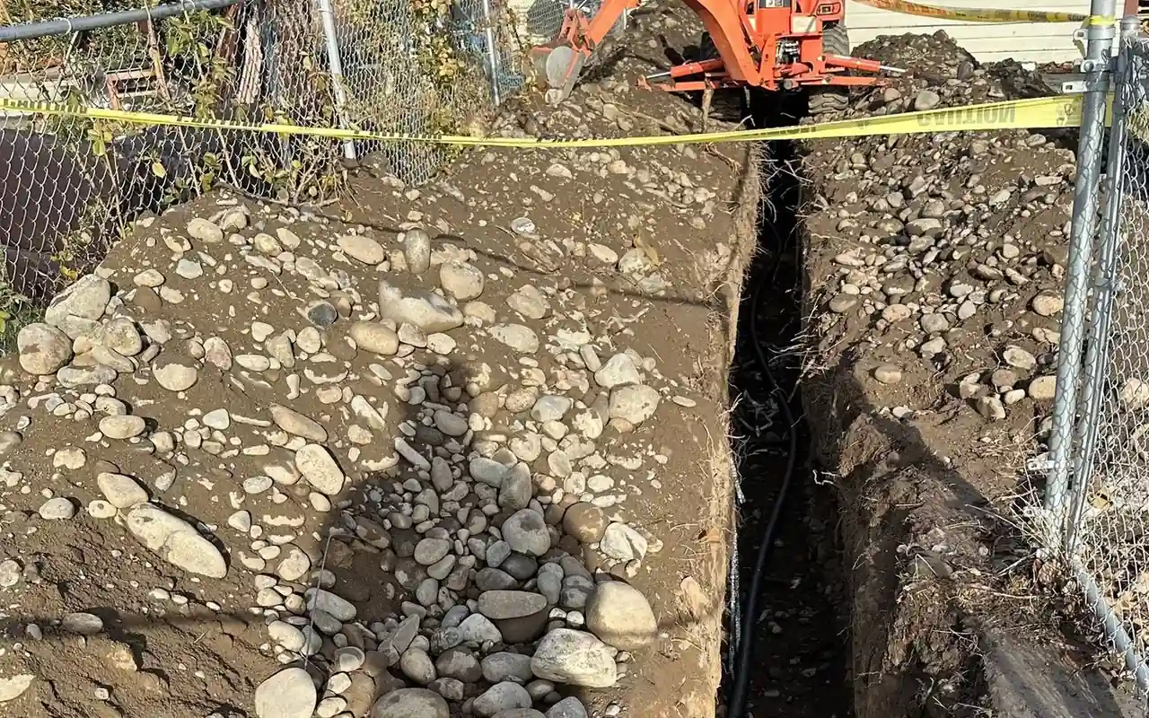 CSR's orange Kubota backhoe digging a water-line trench through Yakima cobble behind a chain-link fence